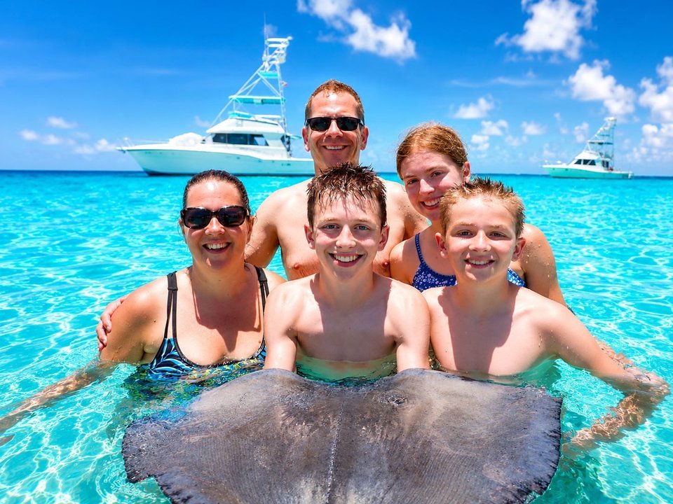 Family of five smiling together in turquoise water beside a southern stingray at Stingray City on a Cayman Adventure Tours excursion in Grand Cayman